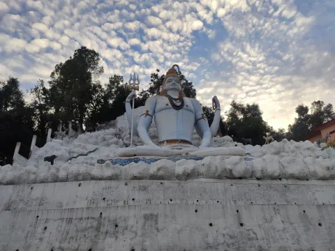 The large, pale blue statue of a meditating Lord Shiva with his trident, set against a dramatic, cloud-filled sky in Ranikhet