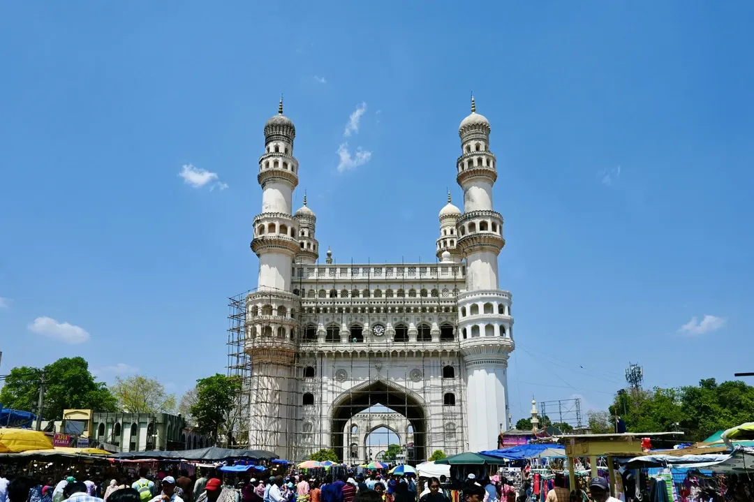 The magnificent Charminar monument, with its four grand minarets, rises above a colorful street market under a clear blue sky