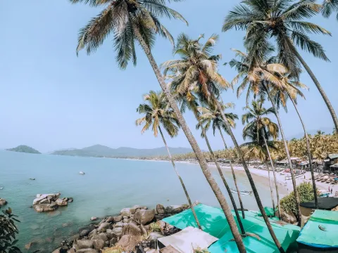 Tropical Goa beach with swaying palm trees, turquoise water, rocky shoreline, and distant beach huts under clear blue skies