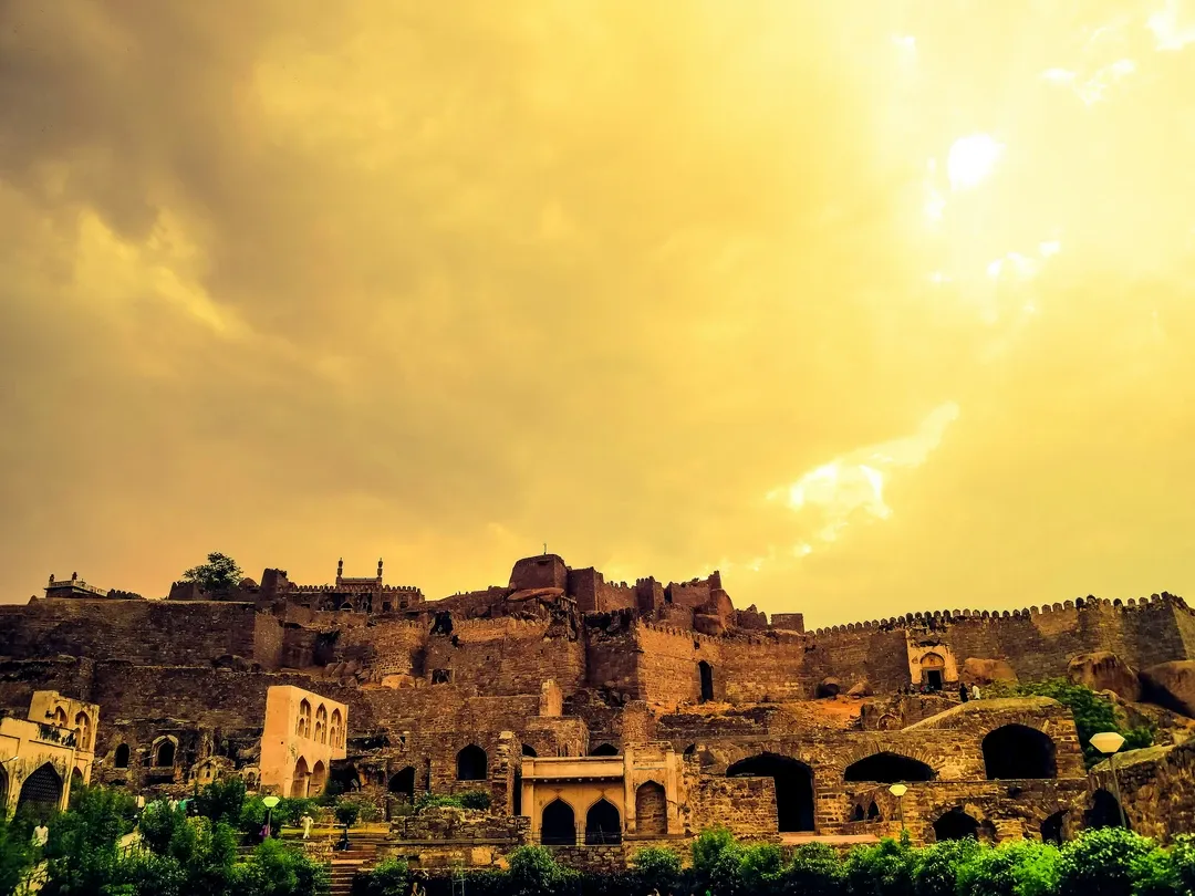 The ancient ruins and imposing stone ramparts of the Golkonda Fort stand on a hilltop under a dramatic, golden-tinted sky