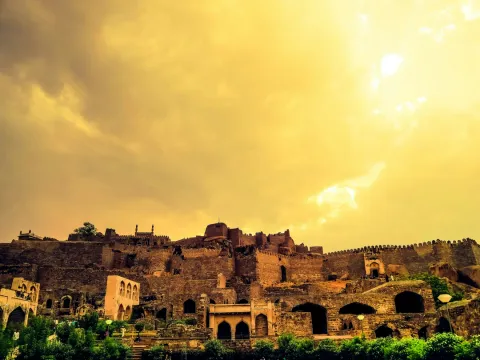 The ancient ruins and imposing stone ramparts of the Golkonda Fort stand on a hilltop under a dramatic, golden-tinted sky