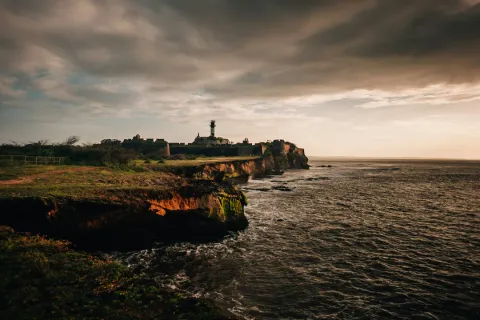 The historic Diu Fort and its lighthouse stand on a rocky cliff overlooking the Arabian Sea under a moody, golden sunset sky