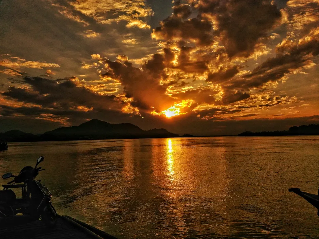 A fiery golden sun sets behind dramatic clouds, its light creating a shimmering reflection on the wide Brahmaputra River in Guwahati