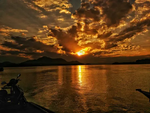 A fiery golden sun sets behind dramatic clouds, its light creating a shimmering reflection on the wide Brahmaputra River in Guwahati