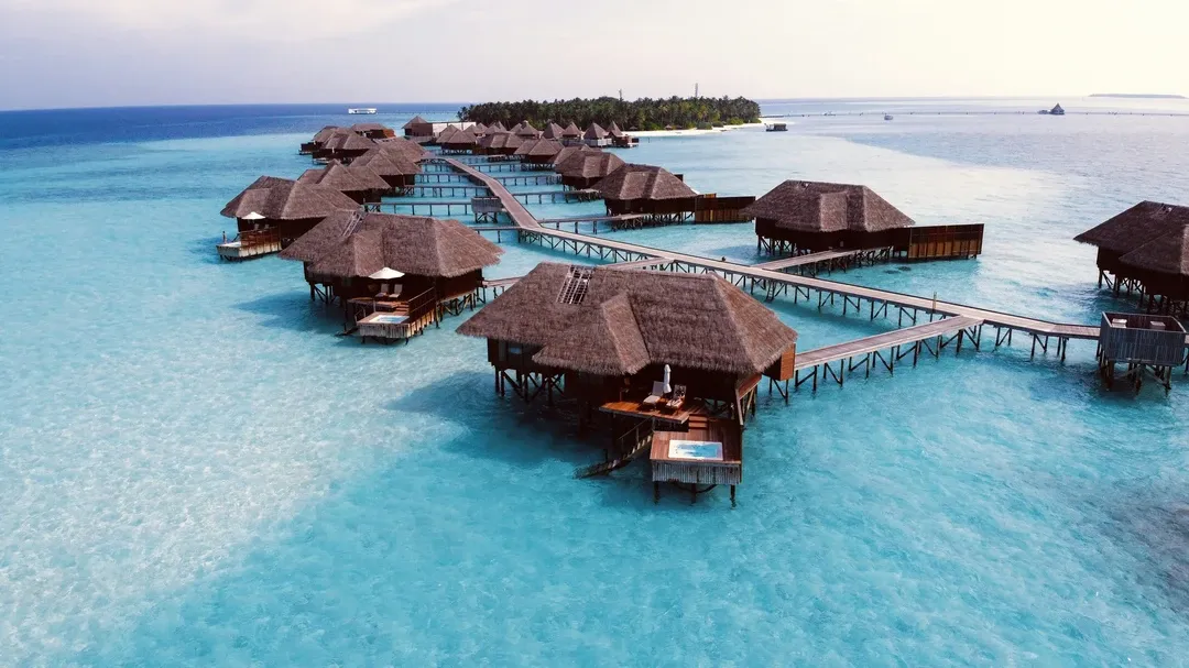 A cluster of thatch-roofed overwater villas connected by wooden jetties over a stunning, crystal-clear turquoise lagoon in the Maldives