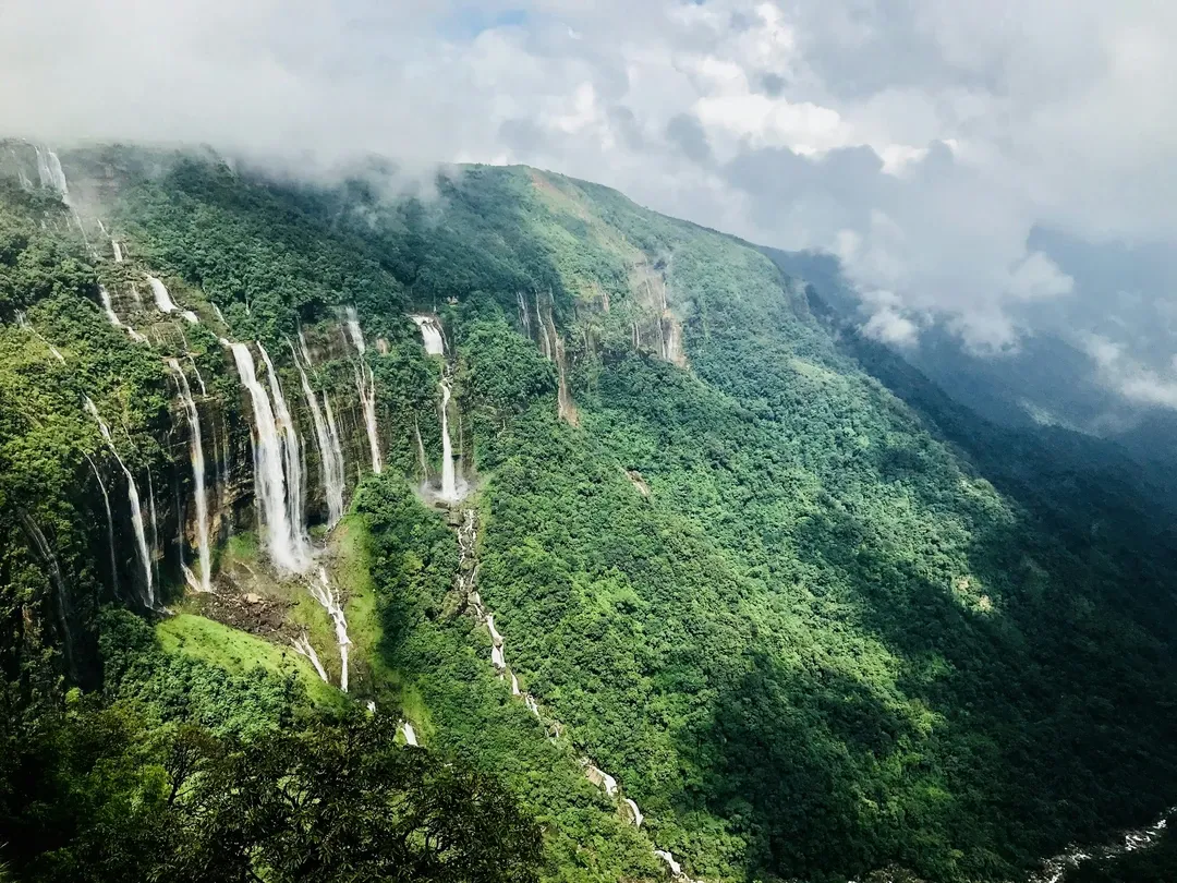 A spectacular monsoon view of the Seven Sisters Waterfalls cascading down a lush green, mist-covered cliff in Cherrapunji