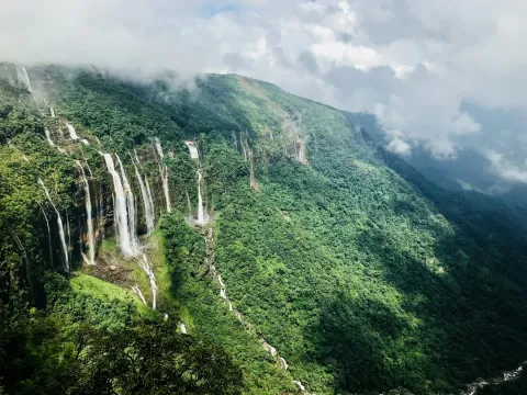 A spectacular monsoon view of the Seven Sisters Waterfalls cascading down a lush green, mist-covered cliff in Cherrapunji