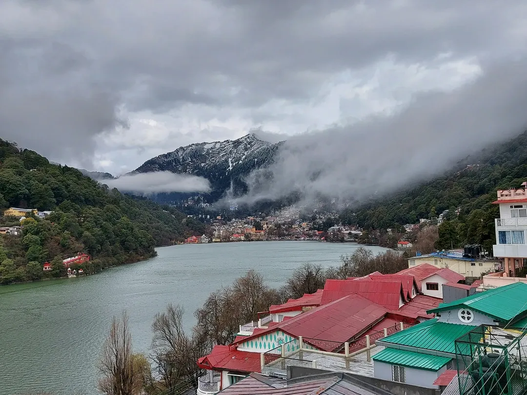 A panoramic view of Naini Lake and the Nainital town, with dramatic clouds rolling over the forested, snow-dusted hills