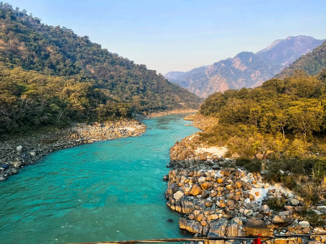 The sacred Ganges River, with its stunning turquoise-colored water, flowing through the rocky, forested hills of Rishikesh