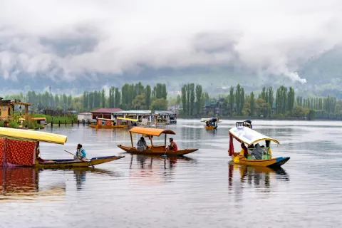 Colorful Shikara boats float on the calm waters of Dal Lake in Srinagar, with houseboats and misty mountains in the background.