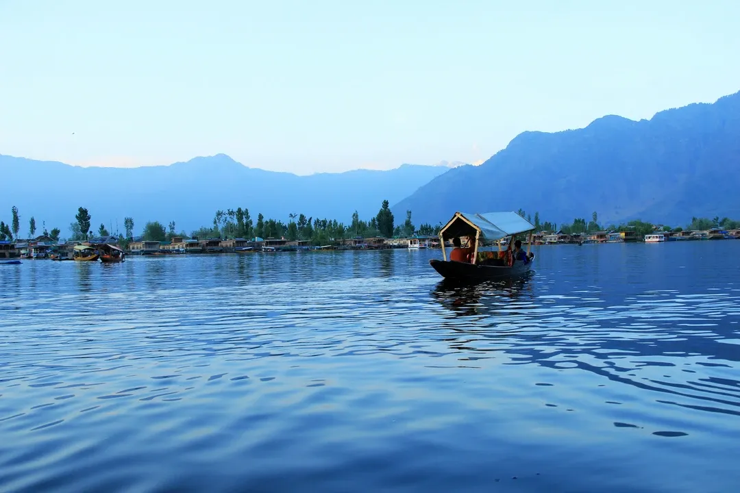 A Shikara boat floats on the calm, blue waters of Dal Lake at dusk, with the silhouettes of mountains in the background