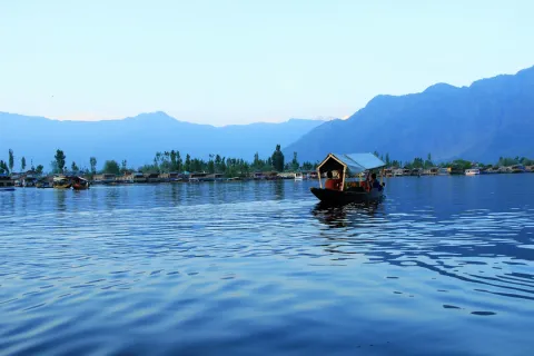 A Shikara boat floats on the calm, blue waters of Dal Lake at dusk, with the silhouettes of mountains in the background