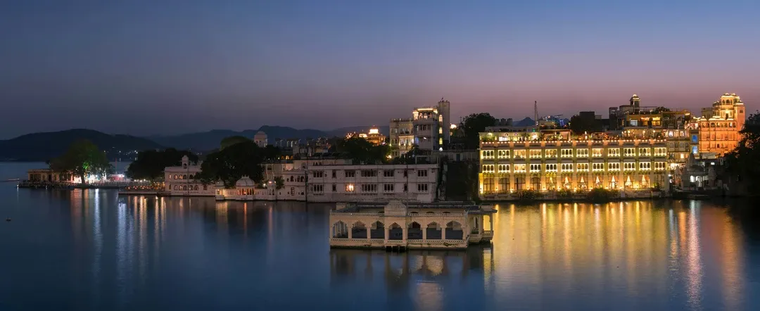 A panoramic view of Udaipur's illuminated palaces and heritage hotels reflecting on the calm waters of Lake Pichola at dusk